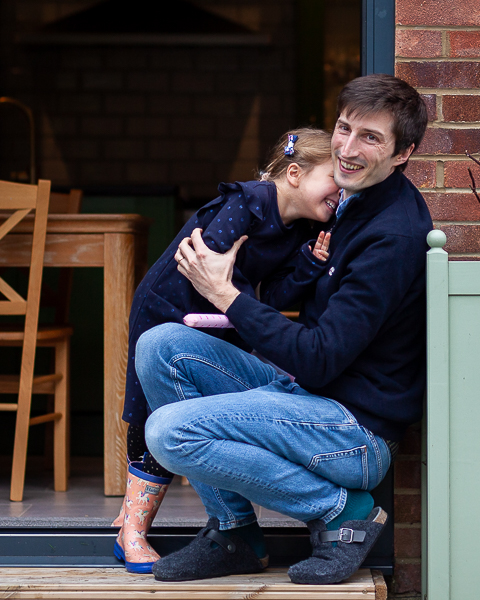 Father crouching by a doorway hugging his daughter, both laughing during a relaxed family session in Fleet Hampshire