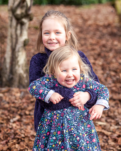 Two sisters hugging and laughing during a natural outdoor family photoshoot in woodland in Hampshire